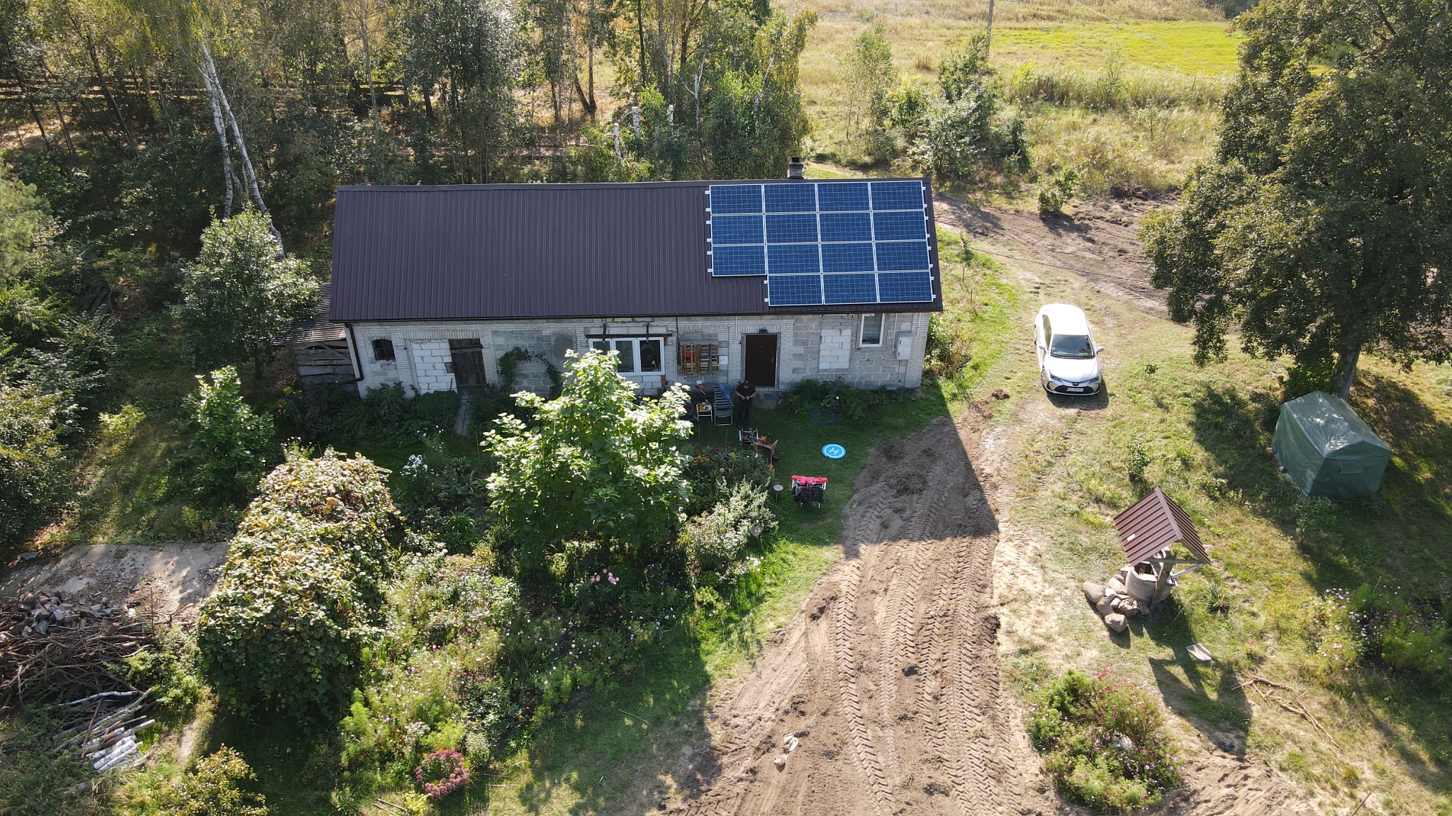Widok z drona na dom z częściowo zamontowanymi panelami fotowoltaicznymi na dachu, z widoczną osobą siedzącą przed domem i białym samochodem zaparkowanym obok