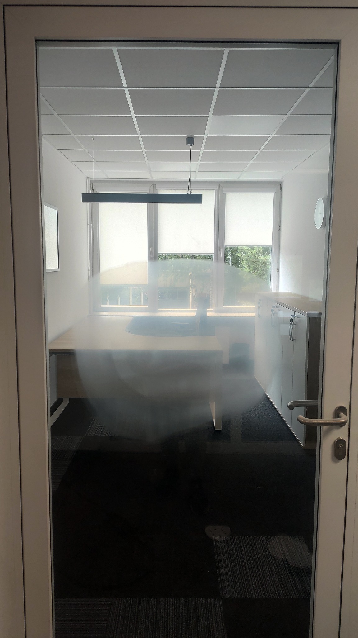 Przez szklane drzwi widok na biuro z biurkiem, szafką i oknami z roletami; widoczne refleksy na szkle.