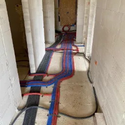 Wnętrze surowego budynku z widocznymi rurami w kolorach czerwonym, niebieskim i czarnym, układanymi na podłodze pomiędzy ścianami z betonu komórkowego; w tle widoczne otwory drzwiowe...