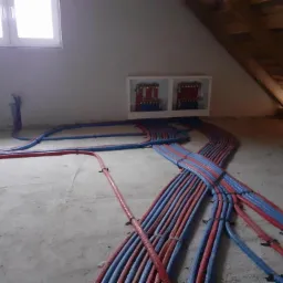Rozłożone na surowym podłożu rurki ogrzewania podłogowego, czerwono-niebieskie, z widoczną skrzynką rozdzielczą na ścianie pod oknem, na poddaszu.