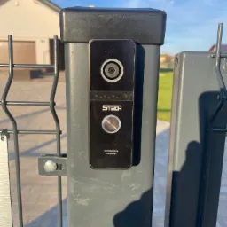 Czarny wideodomofon marki 5TECH zamontowany na szarym słupku ogrodzenia panelowego, widok z bliska na tle domu z garażem.