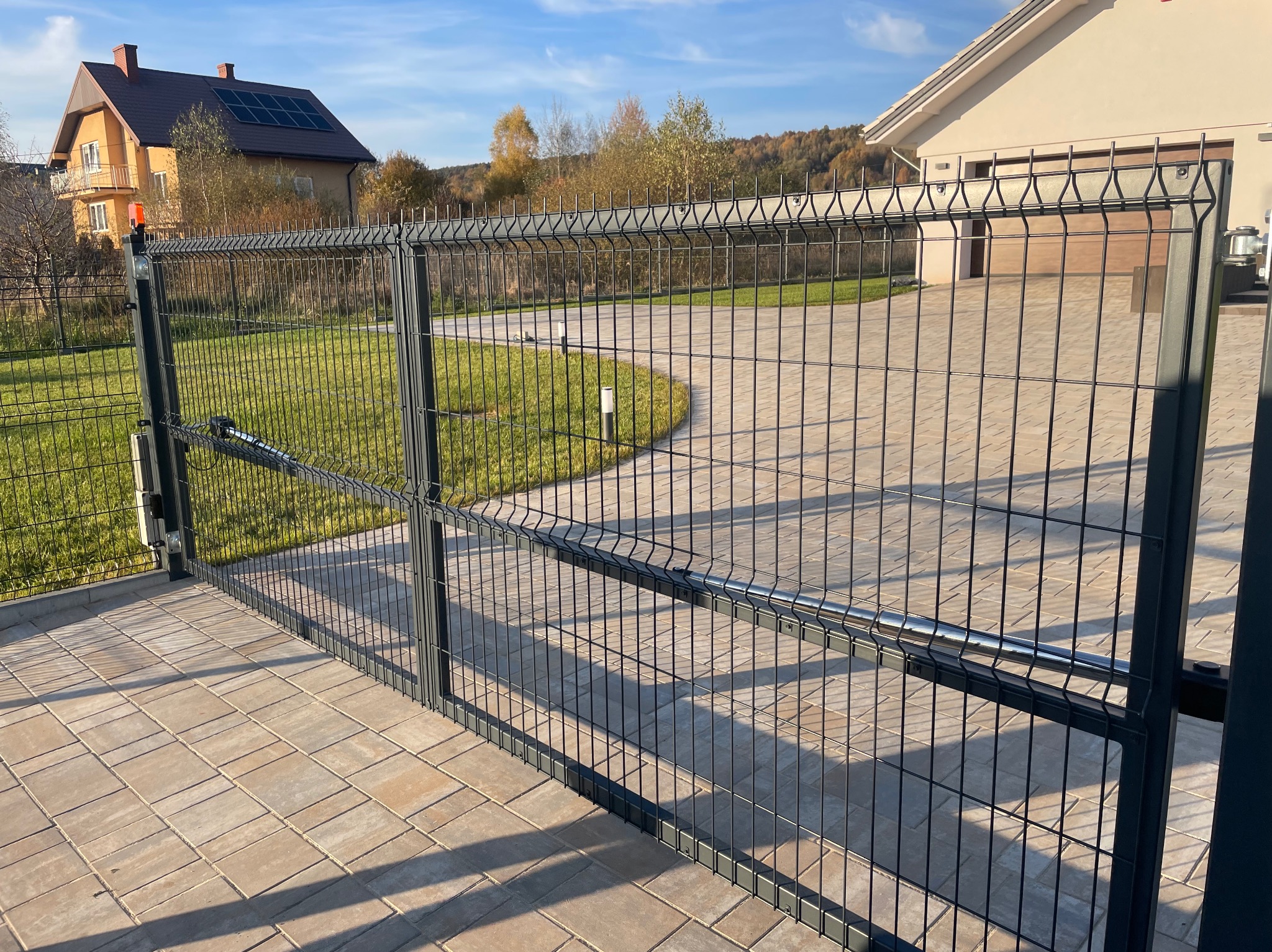 Nowoczesna brama posesyjna z metalowej siatki, automatycznie otwierana, z widocznym mechanizmem napędowym, na tle kostki brukowej i zadbanego trawnika.