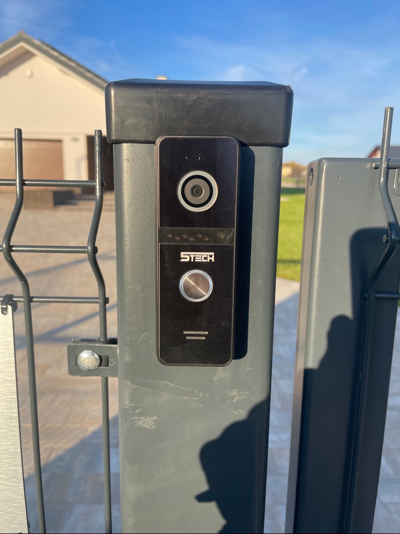 Czarny wideodomofon marki 5TECH zamontowany na szarym słupku ogrodzenia panelowego, widok z bliska na tle domu z garażem.