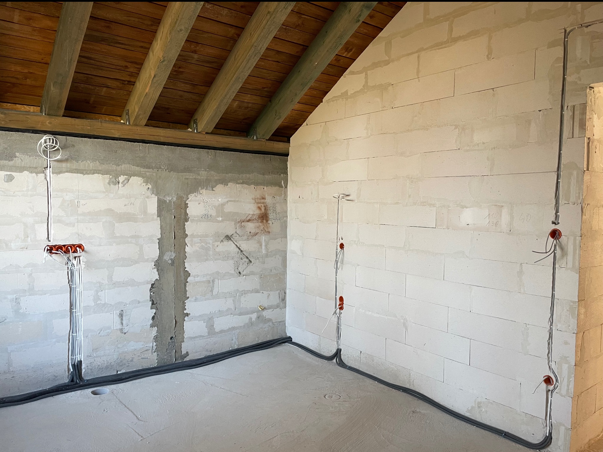 Wnętrze w surowym stanie z widoczną instalacją elektryczną: kable w peszlach prowadzone wzdłuż ścian z bloczków, wystające przewody z puszek. Drewniana konstrukcja dachu.