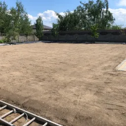 Przygotowany teren pod trawnik z rozłożoną siatką przeciw kretom, widoczne drzewa w tle, drabina w rogu i pracownik z wałem do trawy.