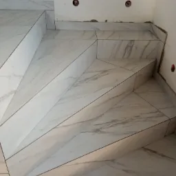 Wyłożone marmuropodobnymi płytkami schody wewnętrzne, widoczne puszki elektryczne w ścianie i ślady po pracach wykończeniowych.