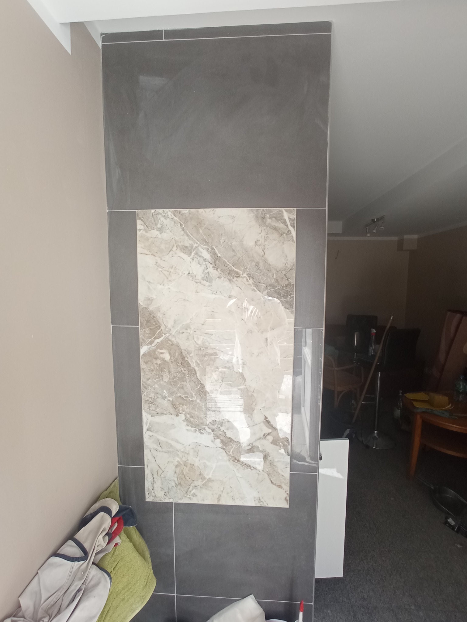 Fragment ściany z szarych i marmurkowych płytek, widoczne niedociągnięcia w postaci zabrudzeń i niedokładności fugowania, w tle umeblowane pomieszczenie.