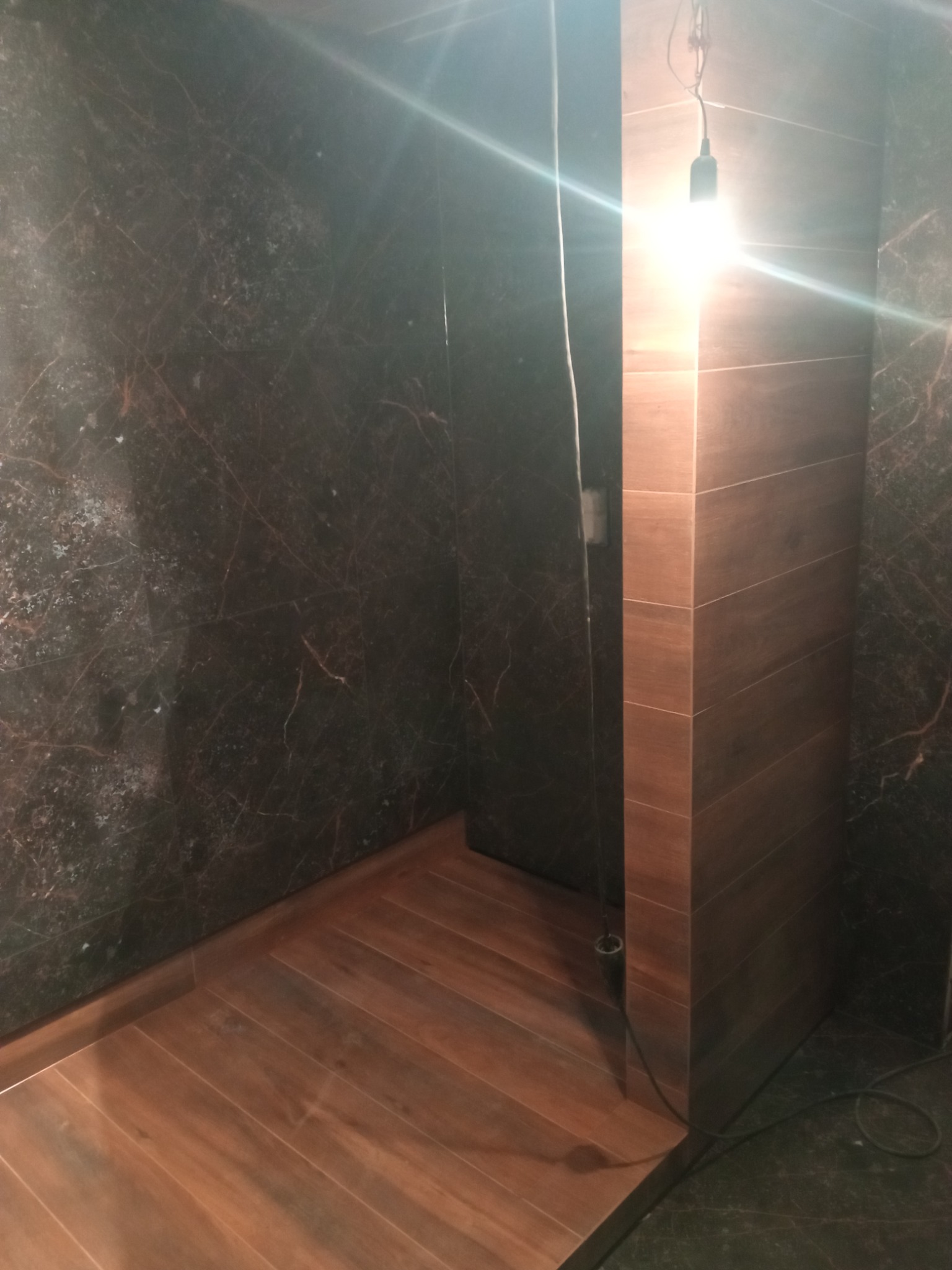 Wykończona łazienka z ciemnymi marmurkowymi płytkami na ścianach i podłodze imitującą drewno, oświetlona wiszącą żarówką.