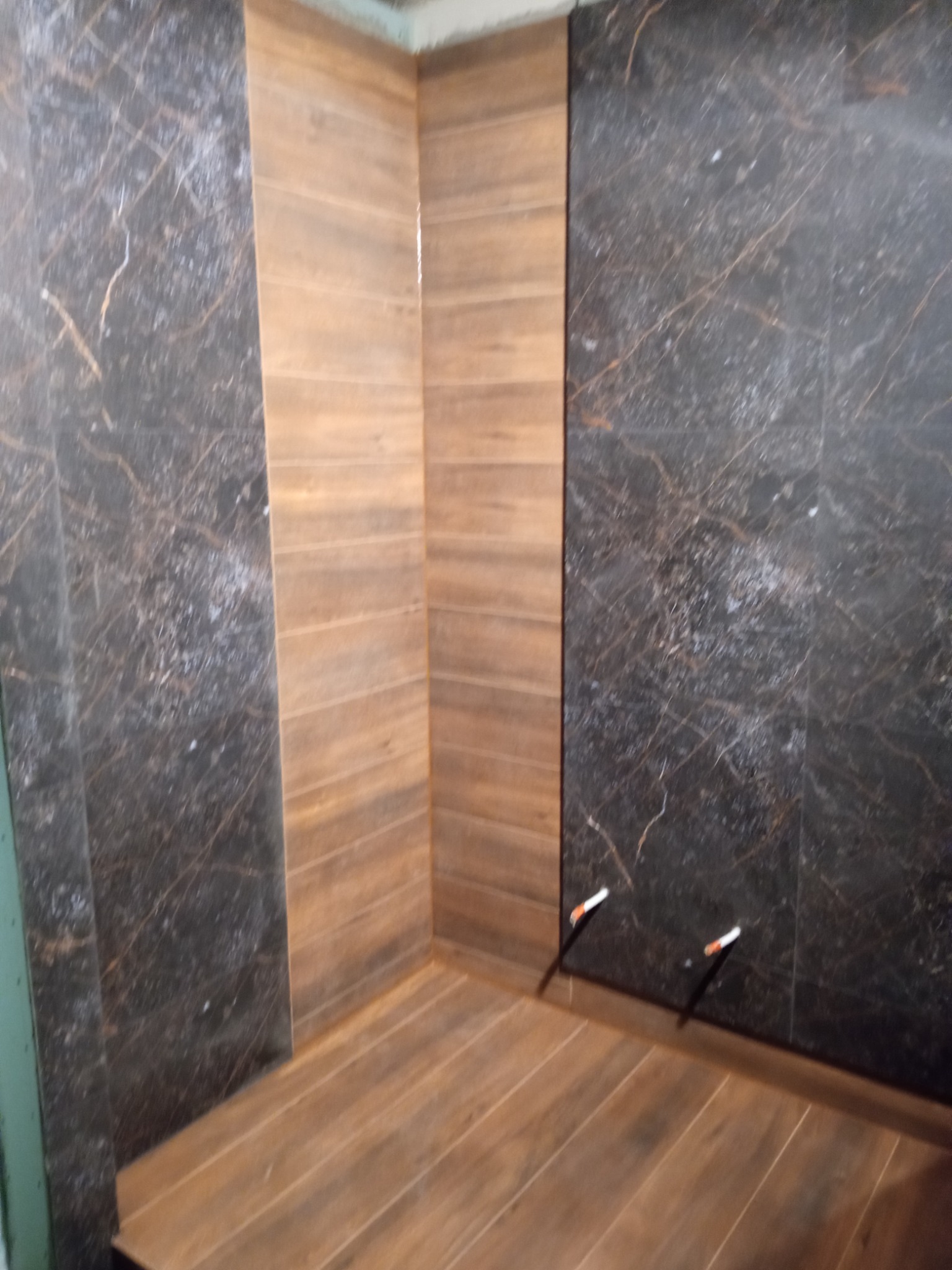 Wykończona kabina prysznicowa z brązowymi płytkami imitującymi drewno na podłodze i ścianie, oraz ciemnymi płytkami z marmurkowym wzorem na ścianach bocznych, widoczne wystające rurki instalacji...