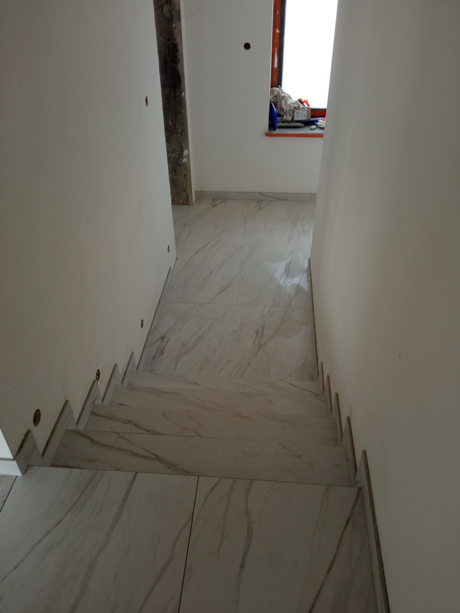Schody wyłożone białymi płytkami imitującymi marmur, prowadzące w dół, z widocznymi fragmentami ścian i okna w tle, w trakcie prac wykończeniowych.
