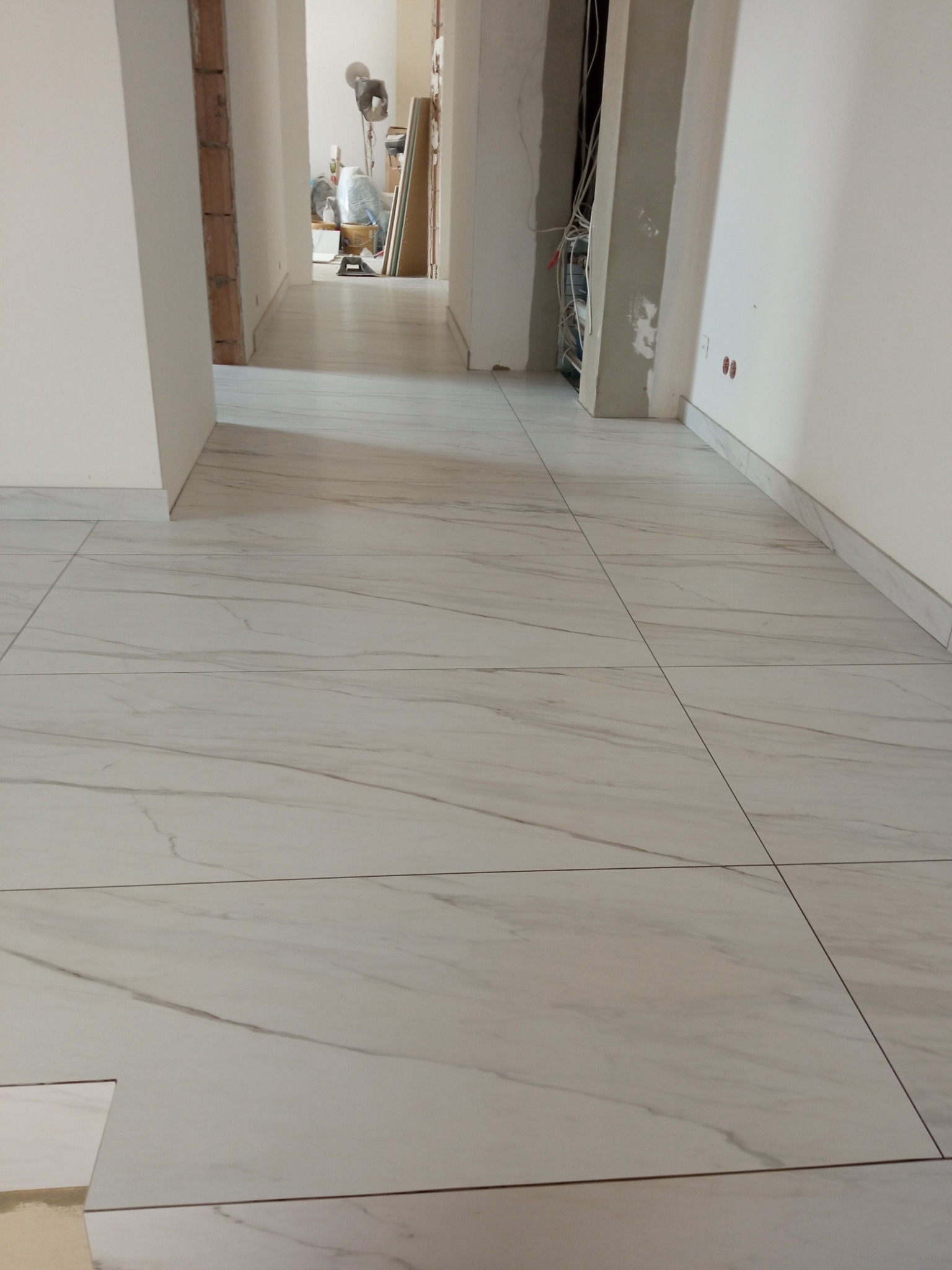 Ułożone białe płytki podłogowe imitujące marmur w korytarzu, widoczne niedokończone prace przy listwach przypodłogowych i brakująca płytka w rogu.