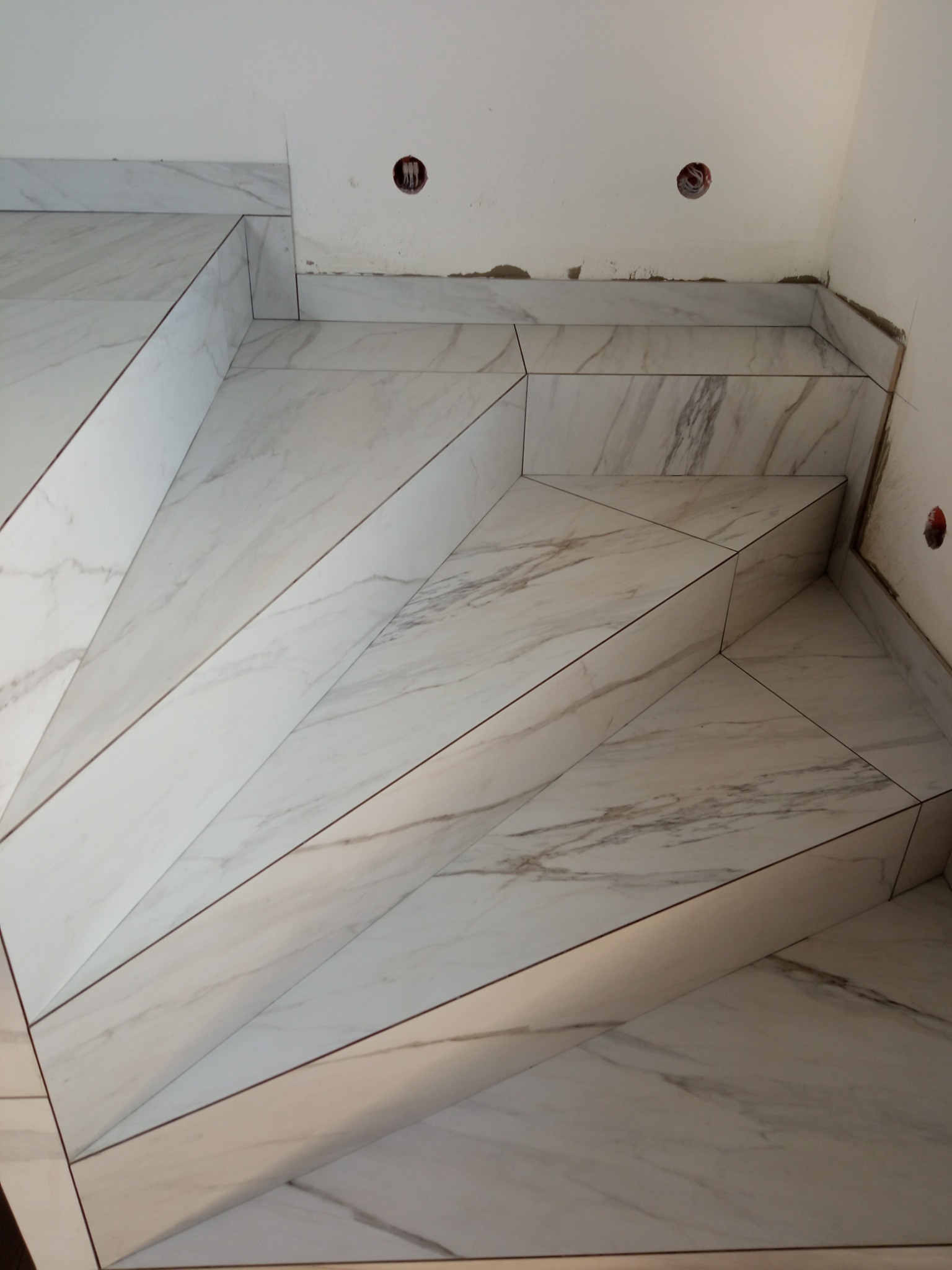 Wyłożone marmuropodobnymi płytkami schody wewnętrzne, widoczne puszki elektryczne w ścianie i ślady po pracach wykończeniowych.