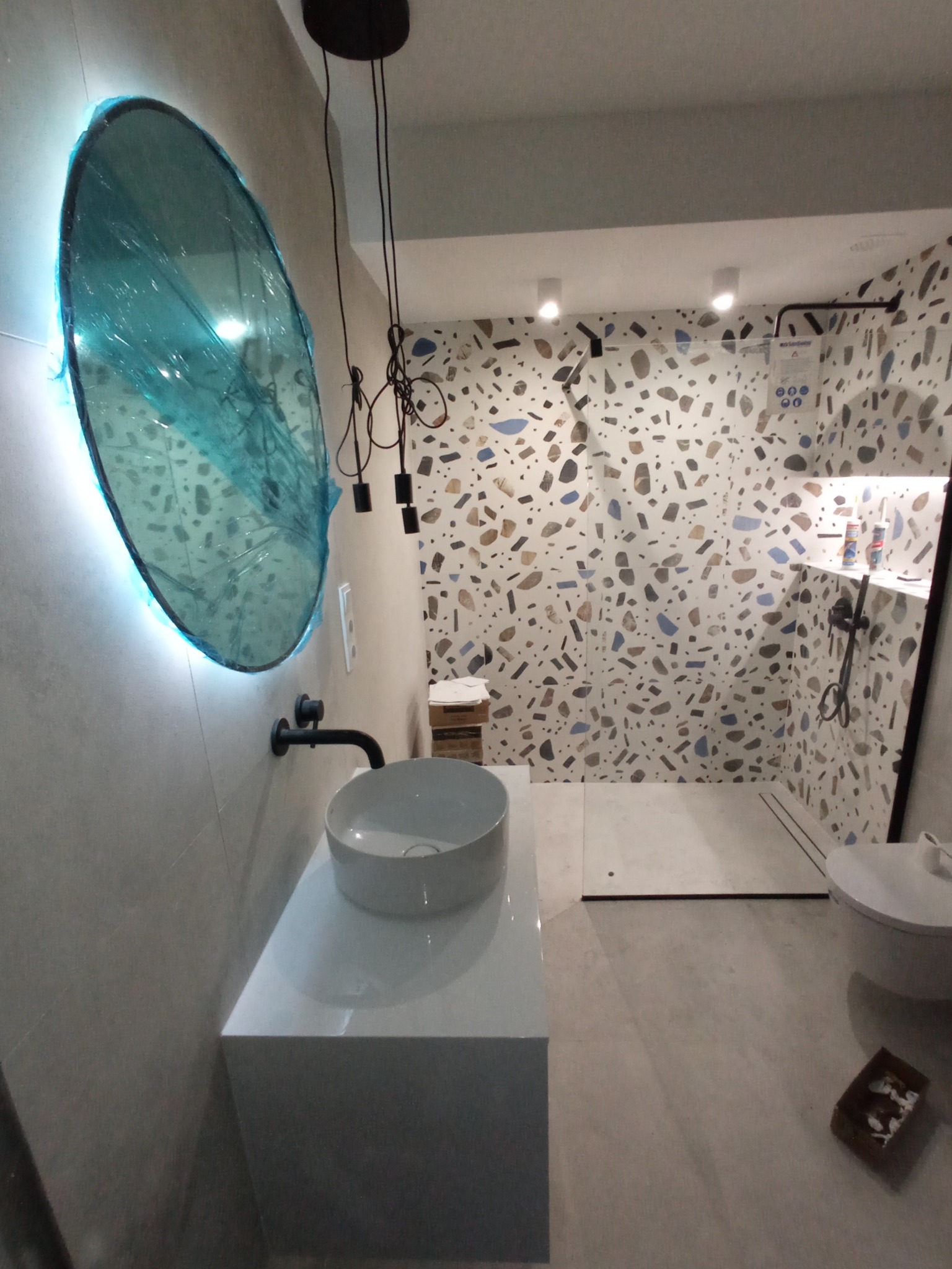 Nowoczesna łazienka z umywalką nablatową, okrągłym lustrem z podświetleniem LED w folii ochronnej, czarną armaturą i mozaikową ścianą w strefie prysznica.