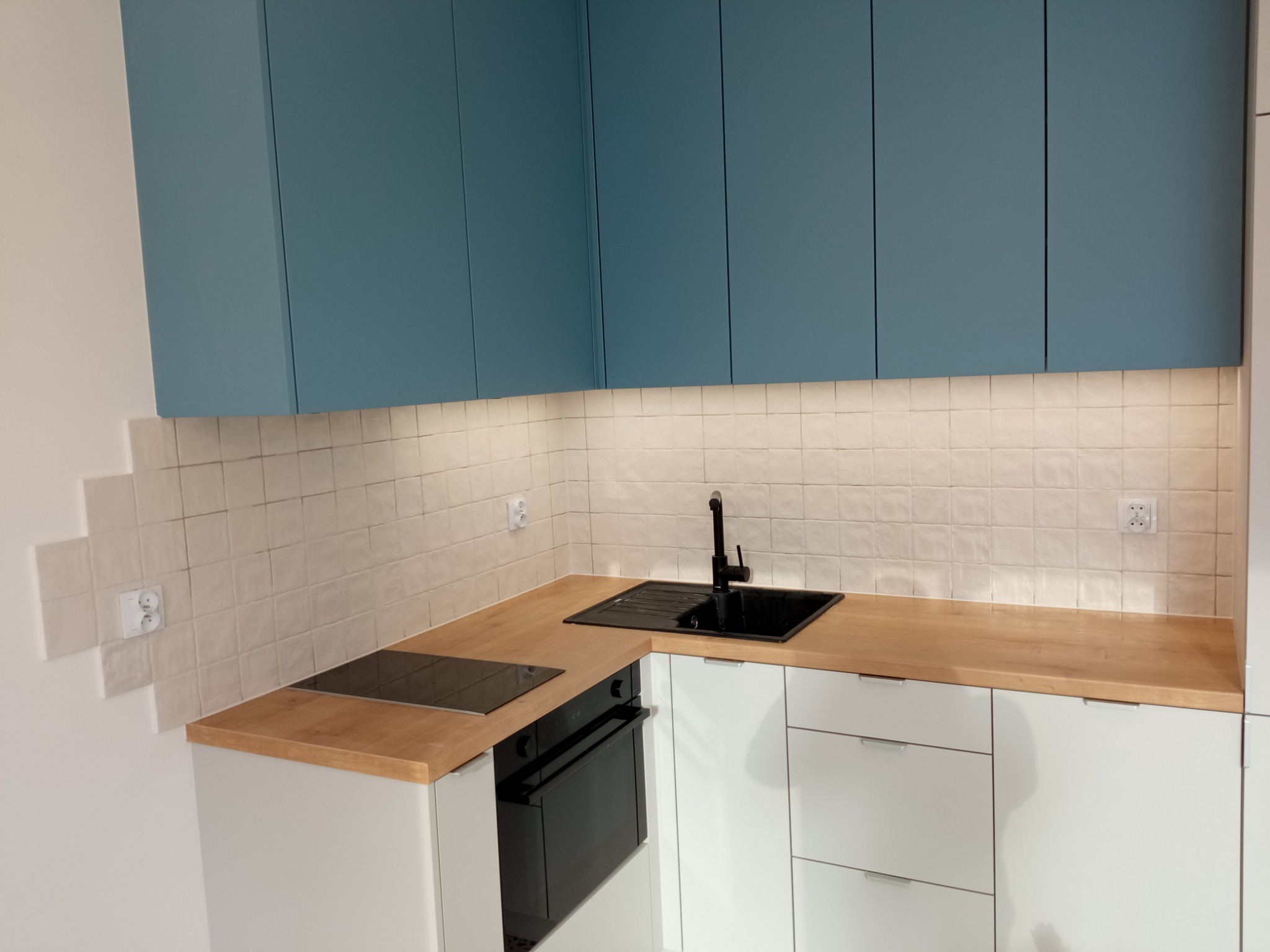 Nowoczesna kuchnia z turkusowymi szafkami górnymi, białymi szafkami dolnymi z poziomymi uchwytami, drewnianym blatem, czarnym zlewem i płytą indukcyjną oraz kremowymi płytkami na ścianie z gniazdkami.