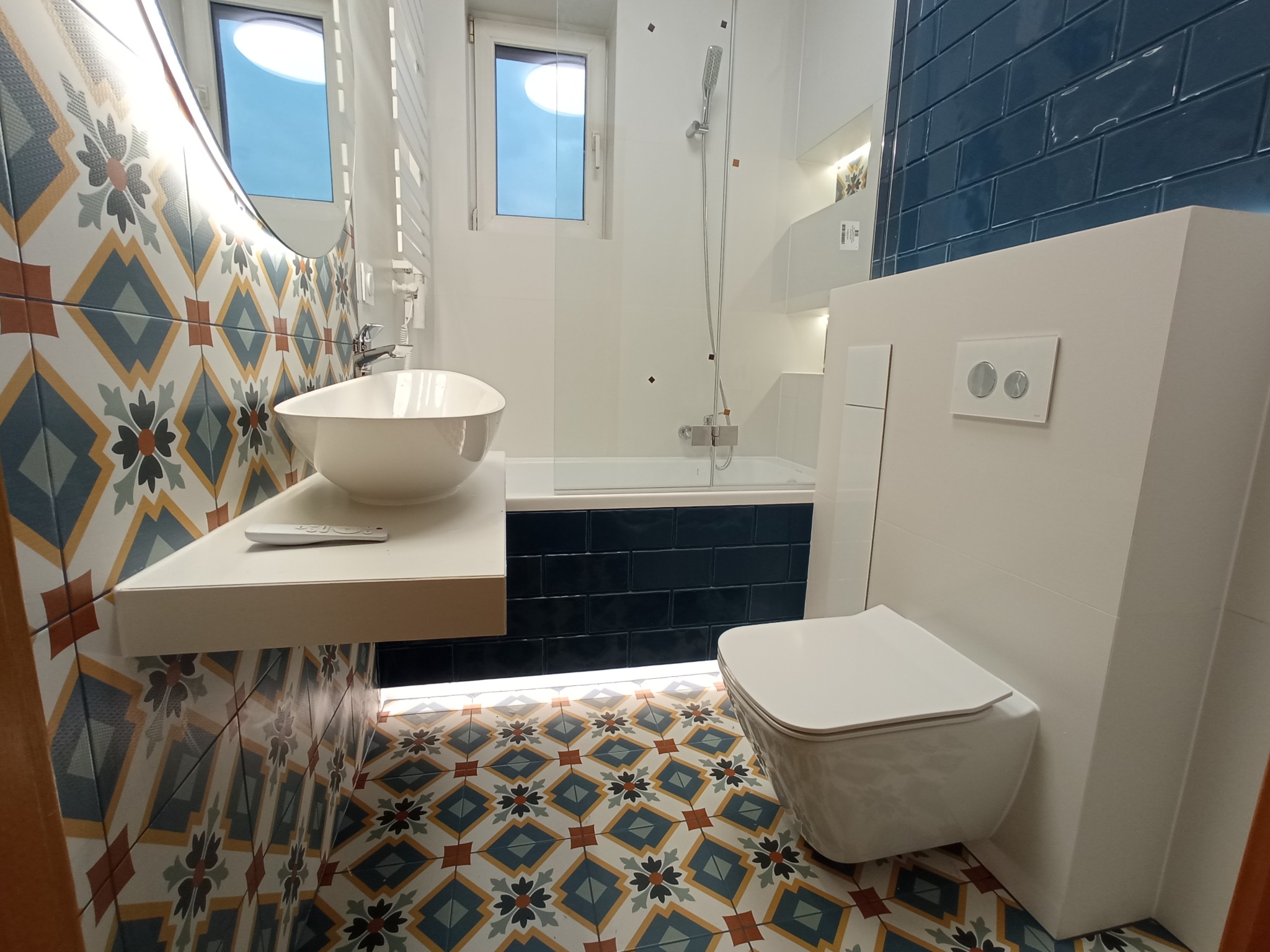 Nowoczesna łazienka z umywalką nablatową, podwieszaną toaletą i wzorzystymi płytkami podłogowymi oraz ściennymi, podświetlenie LED pod szafką umywalkową.