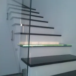 Nowoczesne schody z czarnymi stopniami i białymi podstopnicami, oświetlone zielonymi diodami LED, zabezpieczone szklaną balustradą mocowaną na metalowych uchwytach.