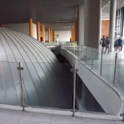 Wnętrze budynku z kopułą pokrytą metalowymi panelami, zabezpieczoną szklaną balustradą z metalowymi słupkami; w tle widoczni ludzie i kolumny.