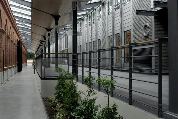 Długa, zewnętrzna galeria z metalowymi poręczami z drewnianym elementem, widok z perspektywy alei z zielonymi krzewami, budynki z cegły i szarego kamienia w tle.