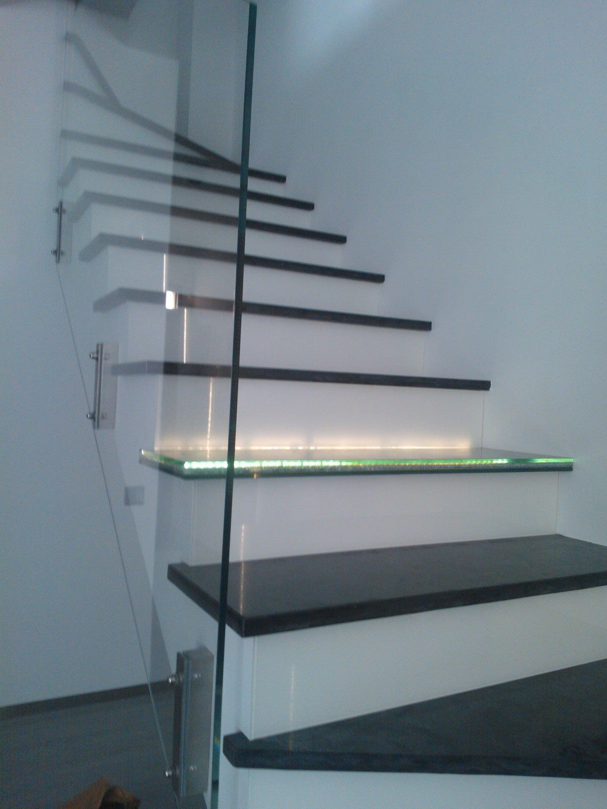 Nowoczesne schody z czarnymi stopniami i białymi podstopnicami, oświetlone zielonymi diodami LED, zabezpieczone szklaną balustradą mocowaną na metalowych uchwytach.
