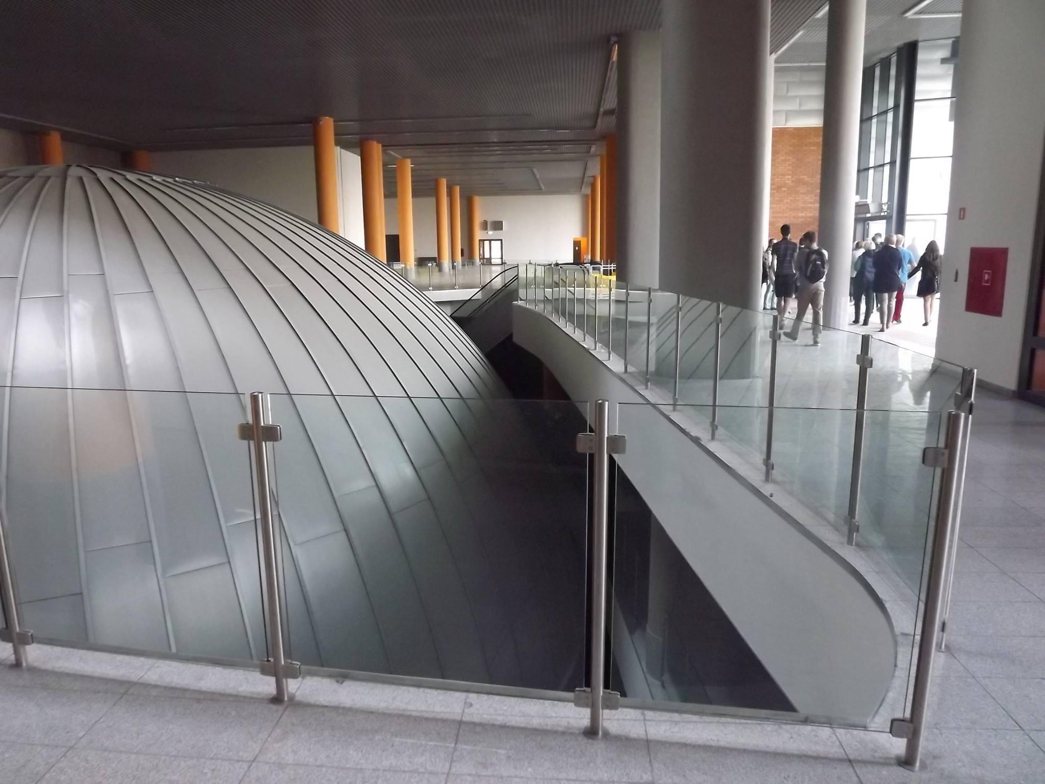 Wnętrze budynku z kopułą pokrytą metalowymi panelami, zabezpieczoną szklaną balustradą z metalowymi słupkami; w tle widoczni ludzie i kolumny.