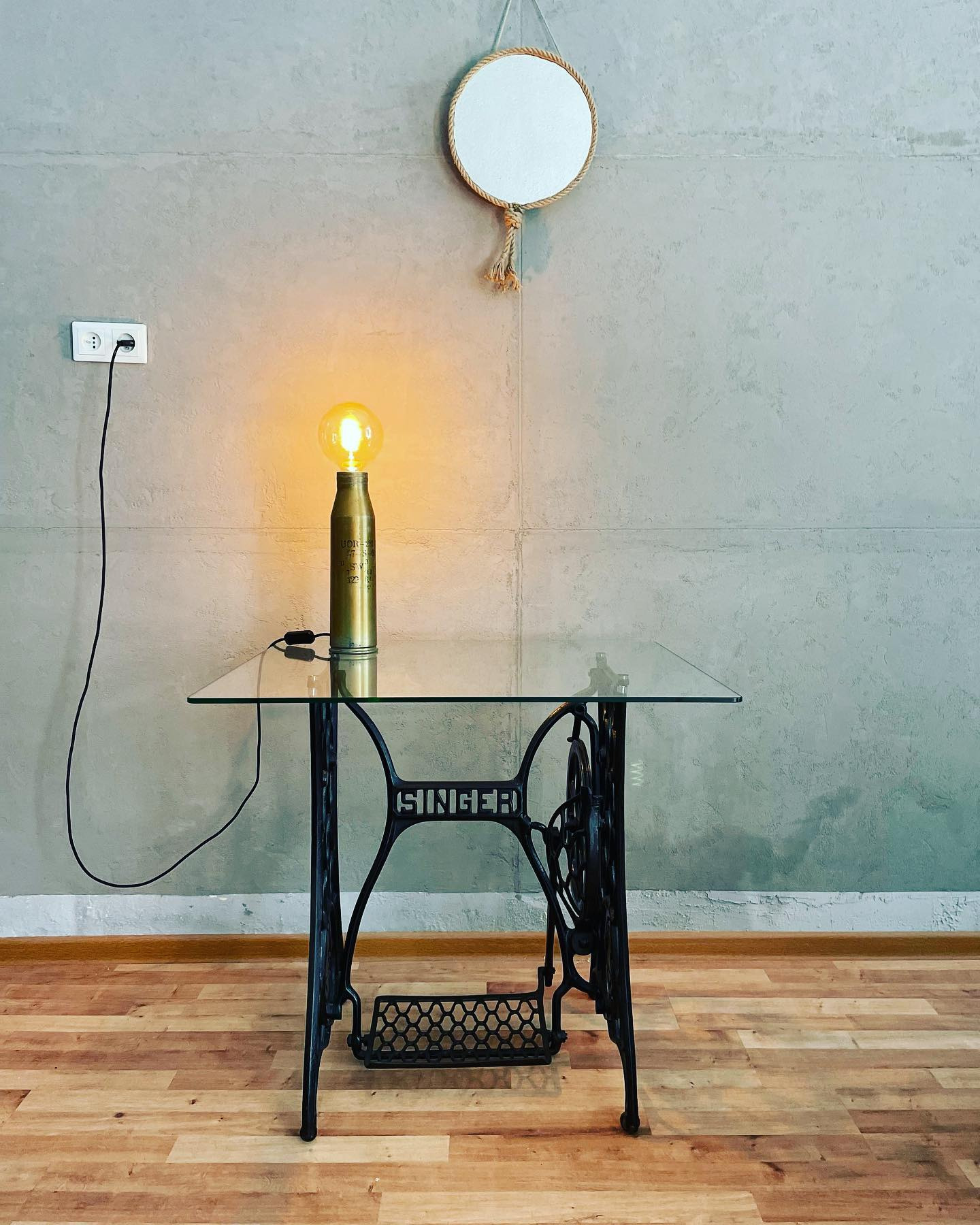Lampa w stylu industrialnym wykonana z łuski pocisku, stojąca na szklanym blacie osadzonym na podstawie starej maszyny do szycia Singer, wiszące lustro z jutowym sznurem w tle.
