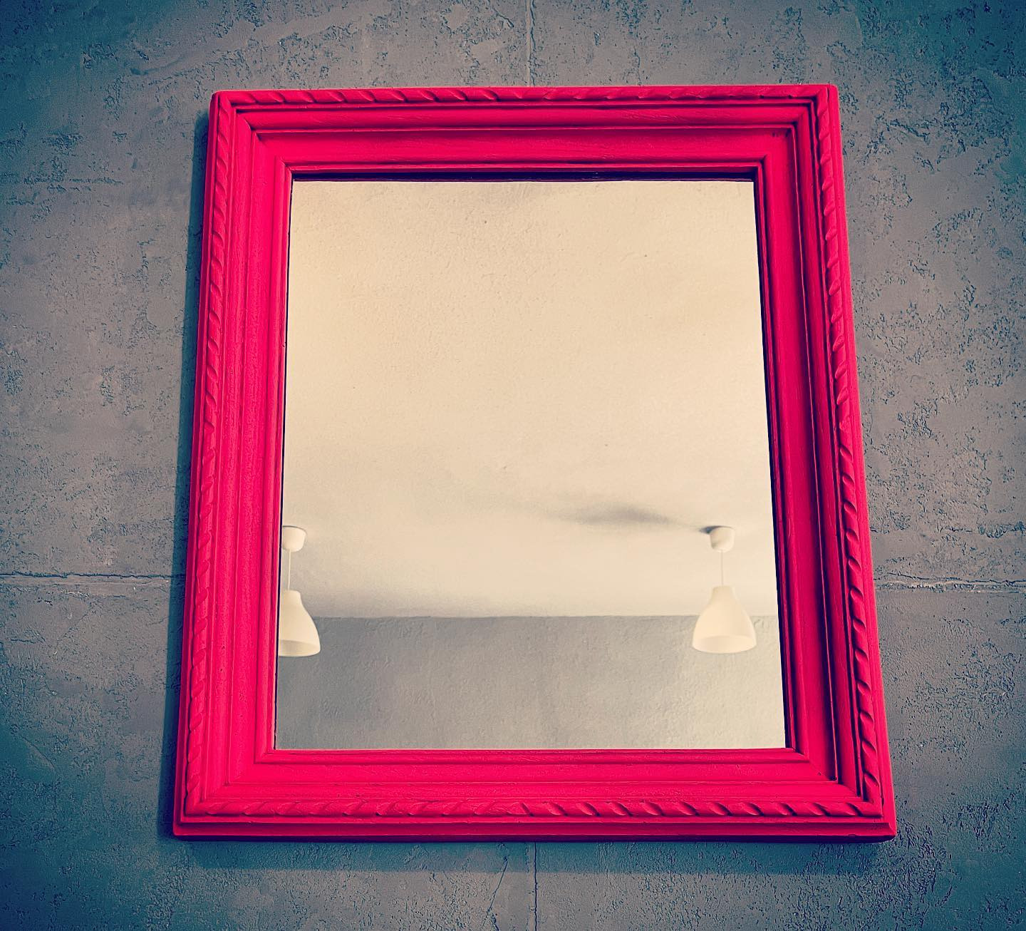 Kwadratowe lustro w jaskrawo różowej, zdobionej ramie zawieszone na szarej, teksturowanej ścianie, odbijające białą lampę sufitową.