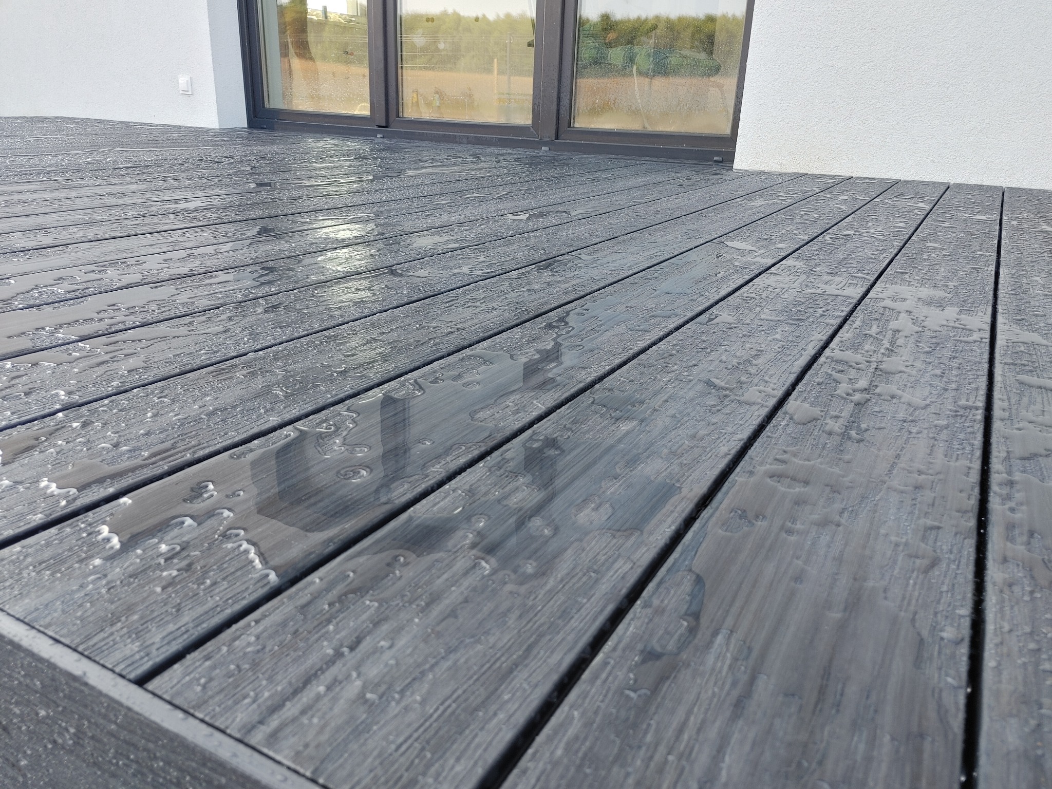 Szare deski tarasowe z widocznymi kroplami wody po deszczu, przylegające do białej ściany z dużymi oknami.