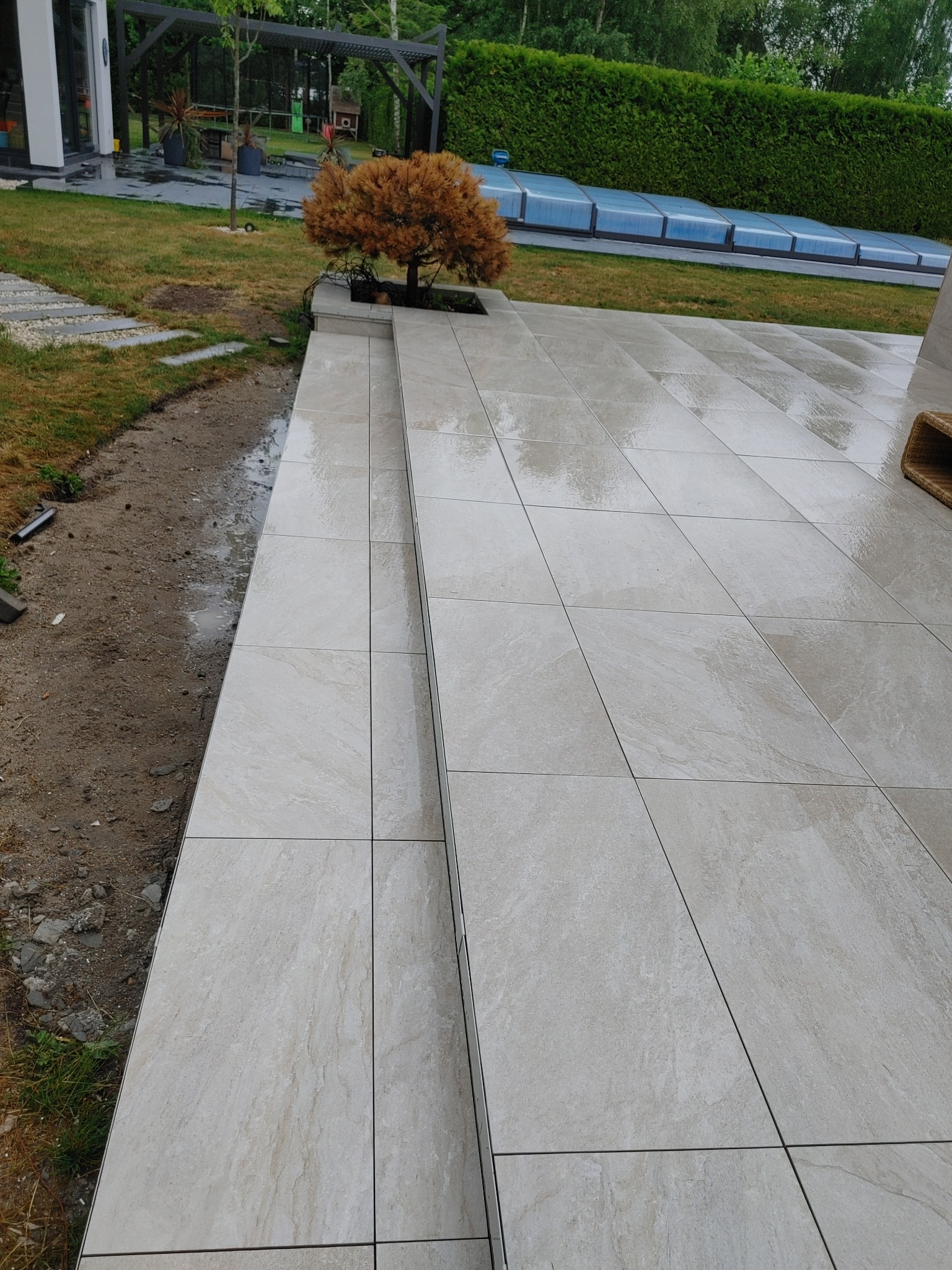 Świeżo ułożony, jasny taras z płytek ceramicznych na zewnątrz, z widoczną krawędzią i stopniami, obok rabaty ziemnej i małego, brązowego drzewka. W tle zarys pergoli i zadaszenia basenu.