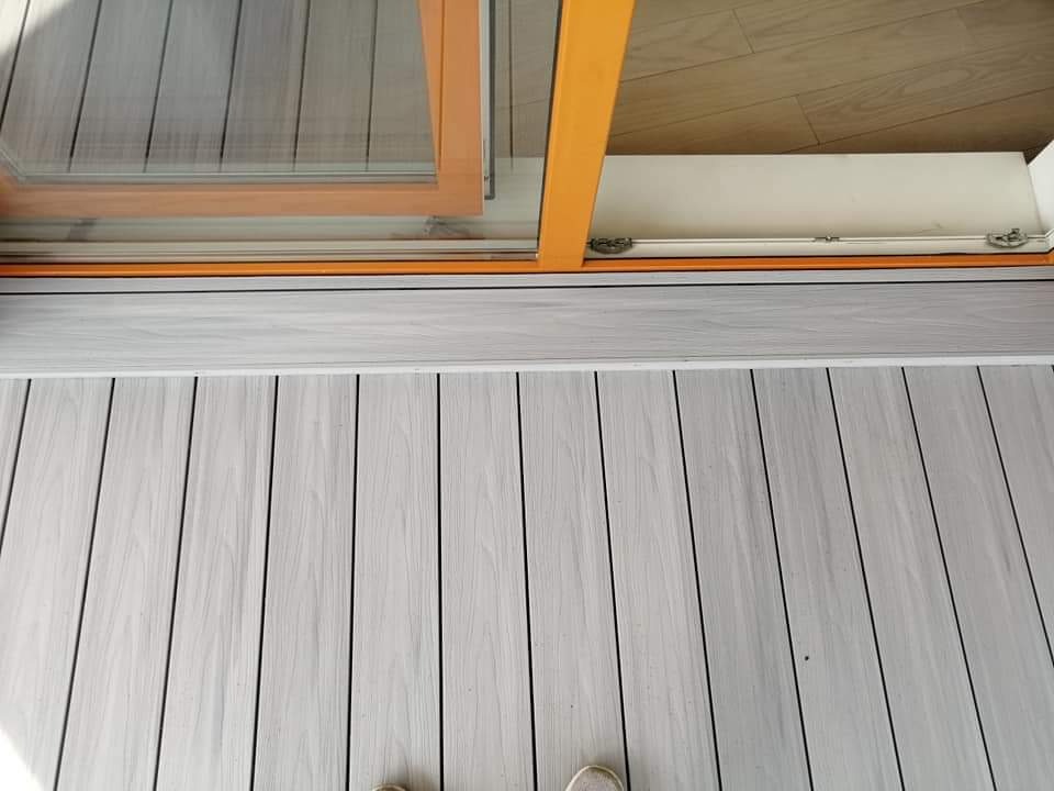 Jasnoszara deska tarasowa ułożona wzdłużnie, widoczna przy progu pomarańczowych drzwi balkonowych z widokiem na parkiet wewnątrz.