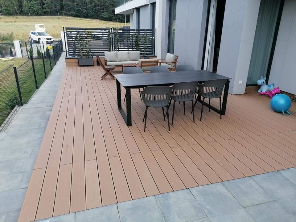 Nowoczesny taras z desek kompozytowych w odcieniach beżu, wyposażony w czarny stół i krzesła, z widokiem na ogród i nowoczesny dom.