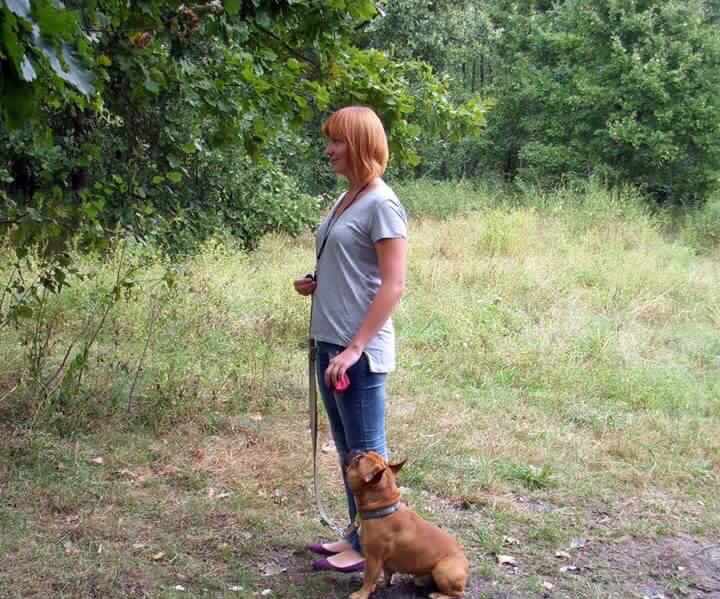 Kobieta z rudymi włosami, w dżinsach i szarej koszulce, trzyma na smyczy brązowego buldoga francuskiego w parku.