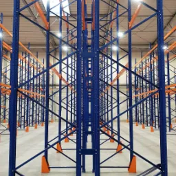 Perspektywa w głąb hali z widokiem na rzędy niebieskich regałów z pomarańczowymi poprzeczkami, oświetlonych światłem jarzeniowym.