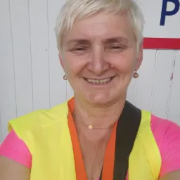 Uśmiechnięta kobieta z krótkimi blond włosami, ubrana w różową koszulkę i żółtą kamizelkę odblaskową z pomarańczowym wykończeniem, pozuje na tle białej ściany z widocznym fragmentem niebieskiej...