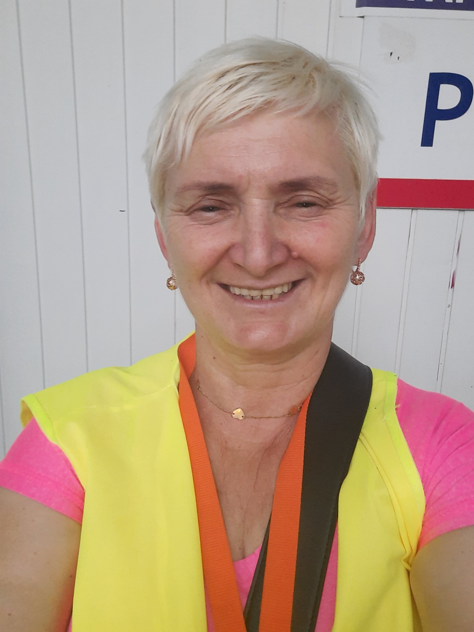 Uśmiechnięta kobieta z krótkimi blond włosami, ubrana w różową koszulkę i żółtą kamizelkę odblaskową z pomarańczowym wykończeniem, pozuje na tle białej ściany z widocznym fragmentem niebieskiej...