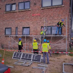 Grupa kursantów w kaskach i kamizelkach odblaskowych montuje rusztowanie przy ceglanym budynku z oknami, na kamizelkach widoczne napisy 'Kursant'.
