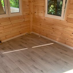 White and Black Co - Wnętrze wykończonego pomieszczenia z drewnianymi ścianami i panelami podłogowymi imitującymi drewno, dwa okna z widokiem na zieleń na zewnątrz.