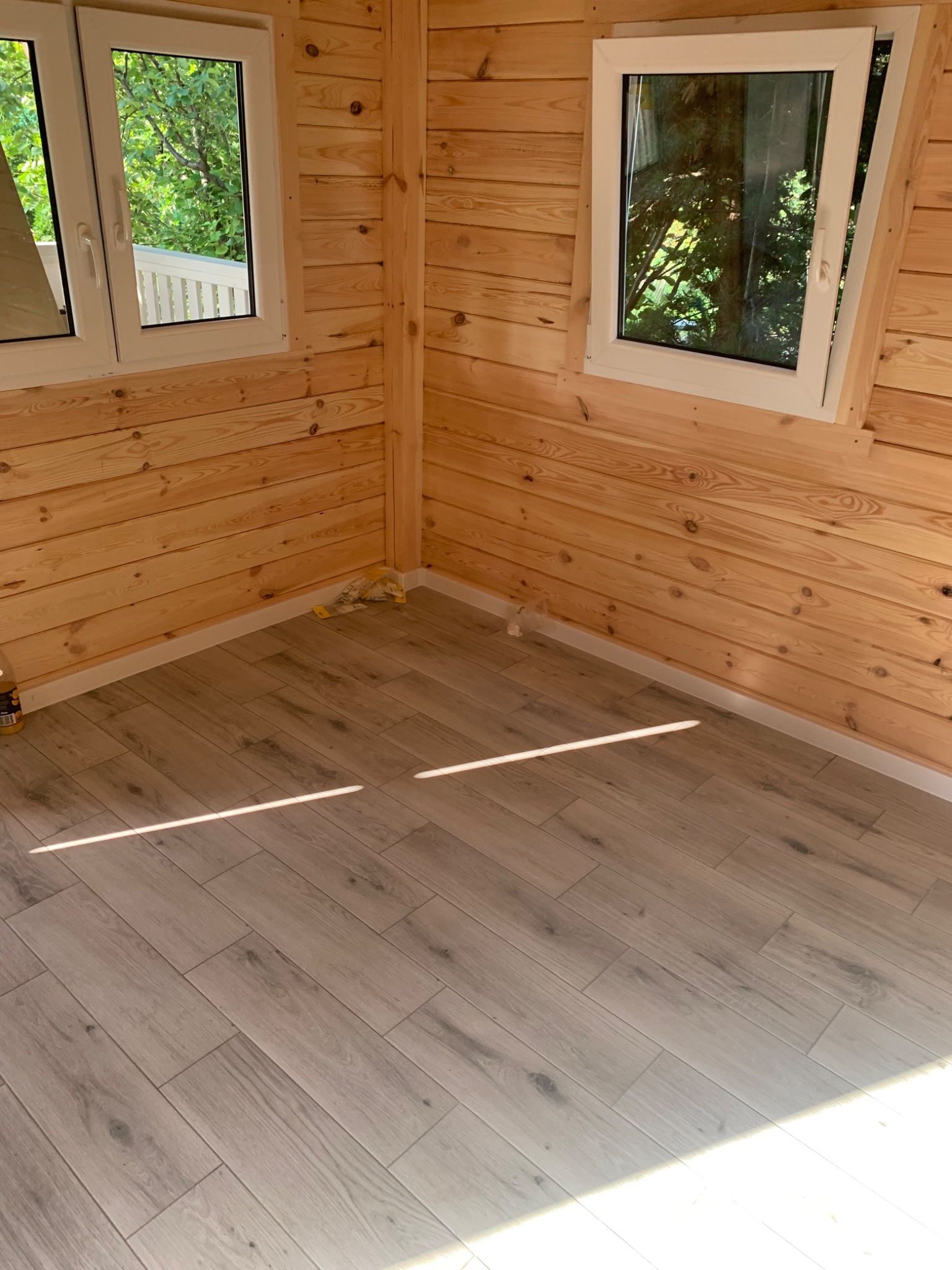 Wnętrze wykończonego pomieszczenia z drewnianymi ścianami i panelami podłogowymi imitującymi drewno, dwa okna z widokiem na zieleń na zewnątrz.