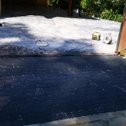 Częściowo wyczyszczona kostka brukowa na podjeździe, widoczne narzędzia do czyszczenia i brama w tle.