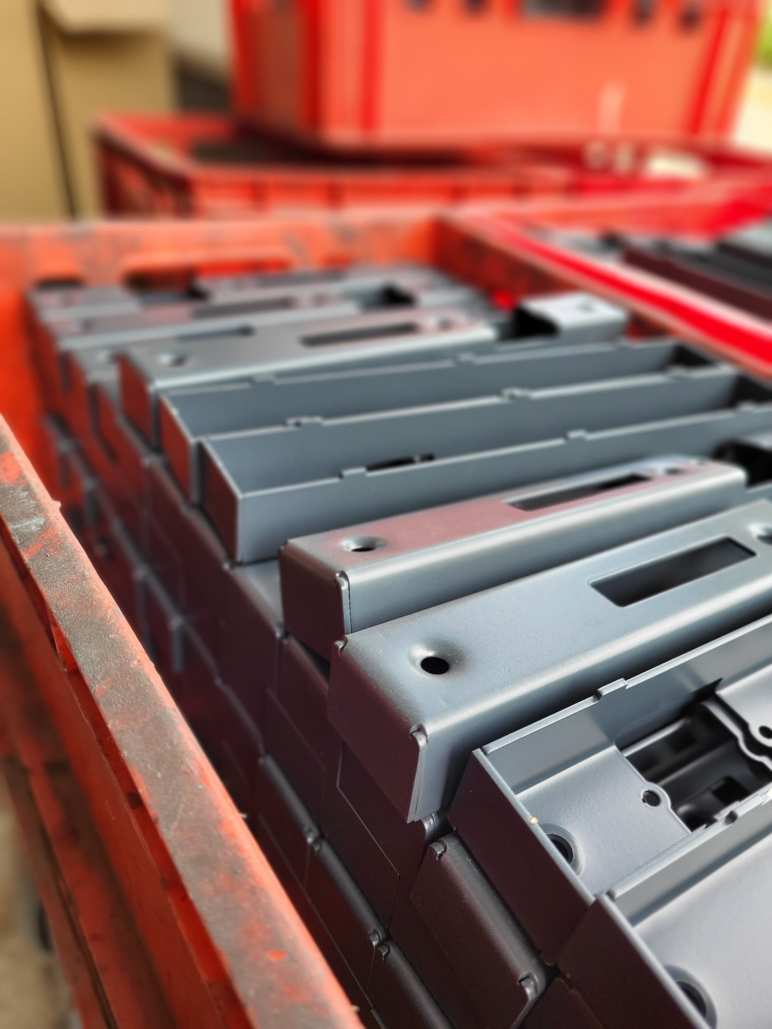 Stos szarych, metalowych obudów z otworami, po malowaniu proszkowym, ułożonych w pomarańczowej skrzynce transportowej.