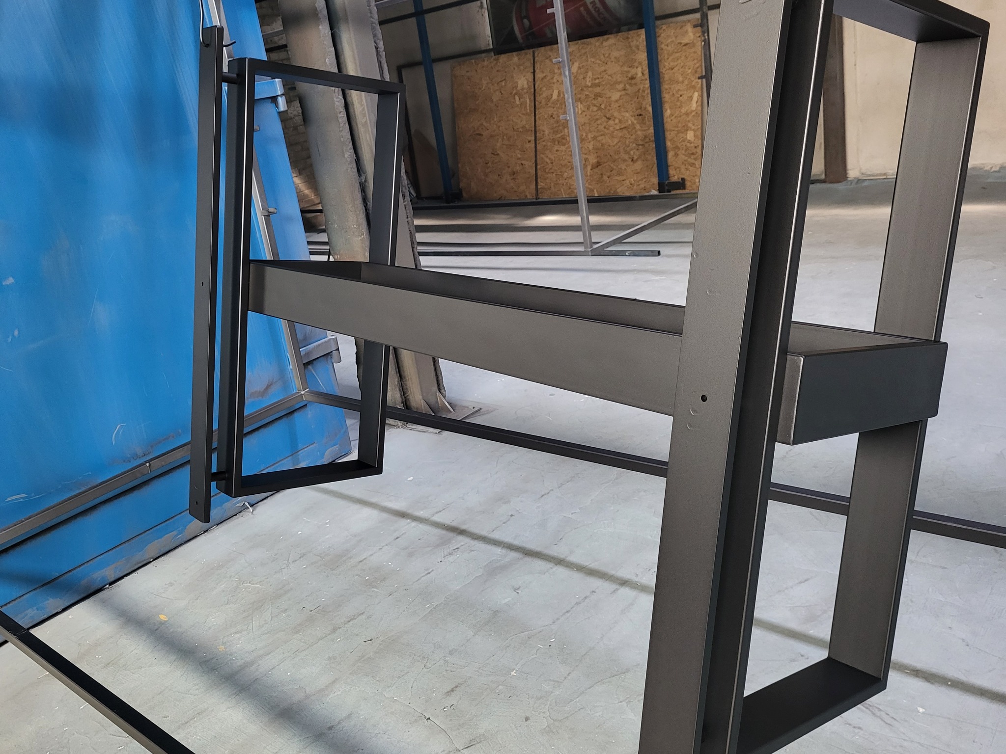 Metalowa konstrukcja krzesła po obróbce, prawdopodobnie pomalowana proszkowo na kolor grafitowy, widoczne elementy ramy i spawów, tło warsztatowe.