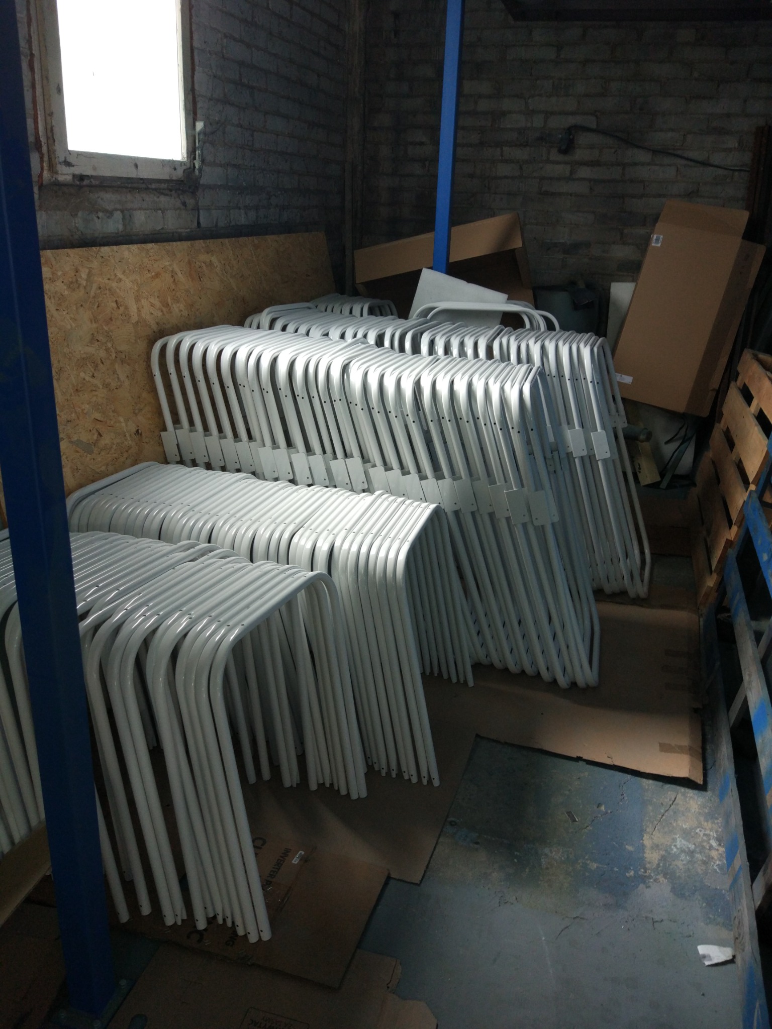 Stosy białych, metalowych stelaży krzeseł po obróbce, ułożone w magazynie na kartonach, widoczne fragmenty ceglanej ściany i okna.