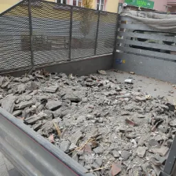 Gruz w metalowej skrzyni samochodu transportowego, widoczne fragmenty cegieł i betonu, w tle budynki miejskie z oknami łukowymi i szyld sklepu Żabka.