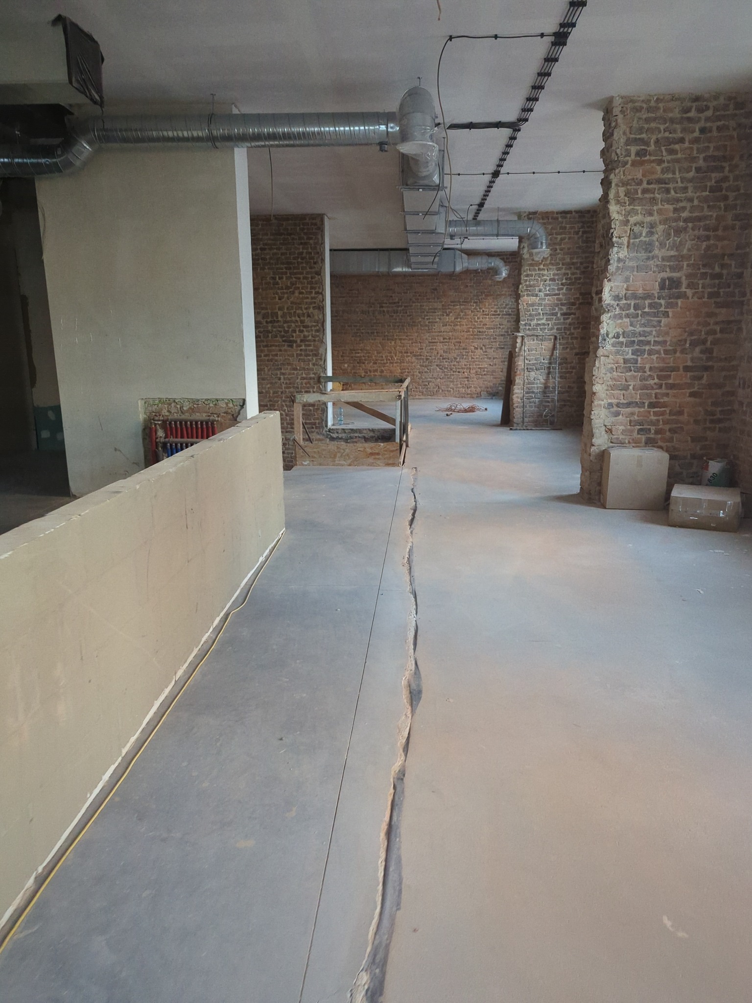 Wnętrze w stanie surowym z widoczną pękniętą posadzką, fragmentami ceglanych ścian, instalacją wentylacyjną pod sufitem i prowizoryczną barierką z desek.