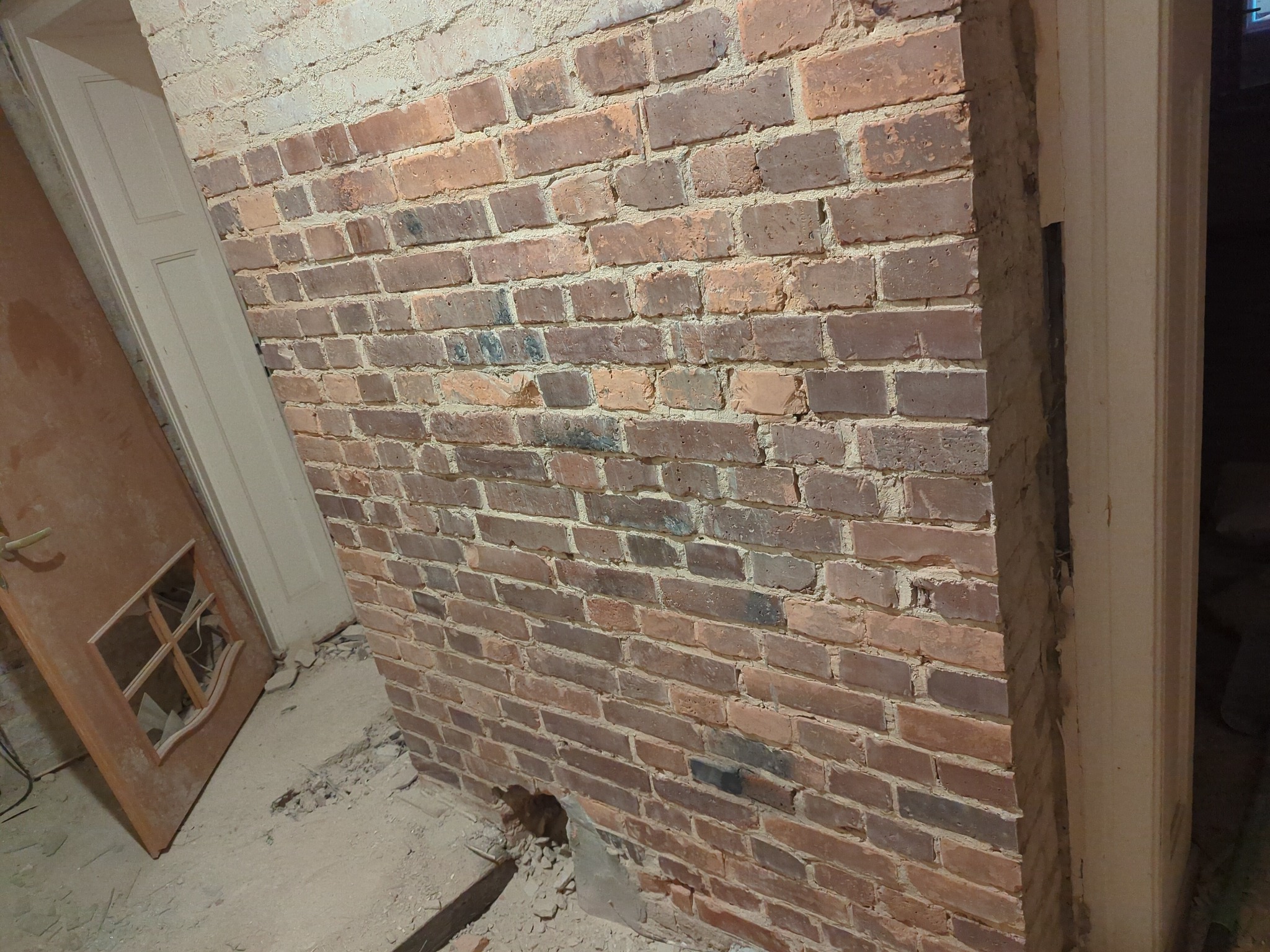 Odsłonięta ceglana ściana wewnętrzna w trakcie remontu, z widocznymi śladami po usuniętych tynkach i elementami stolarki drzwiowej w tle.