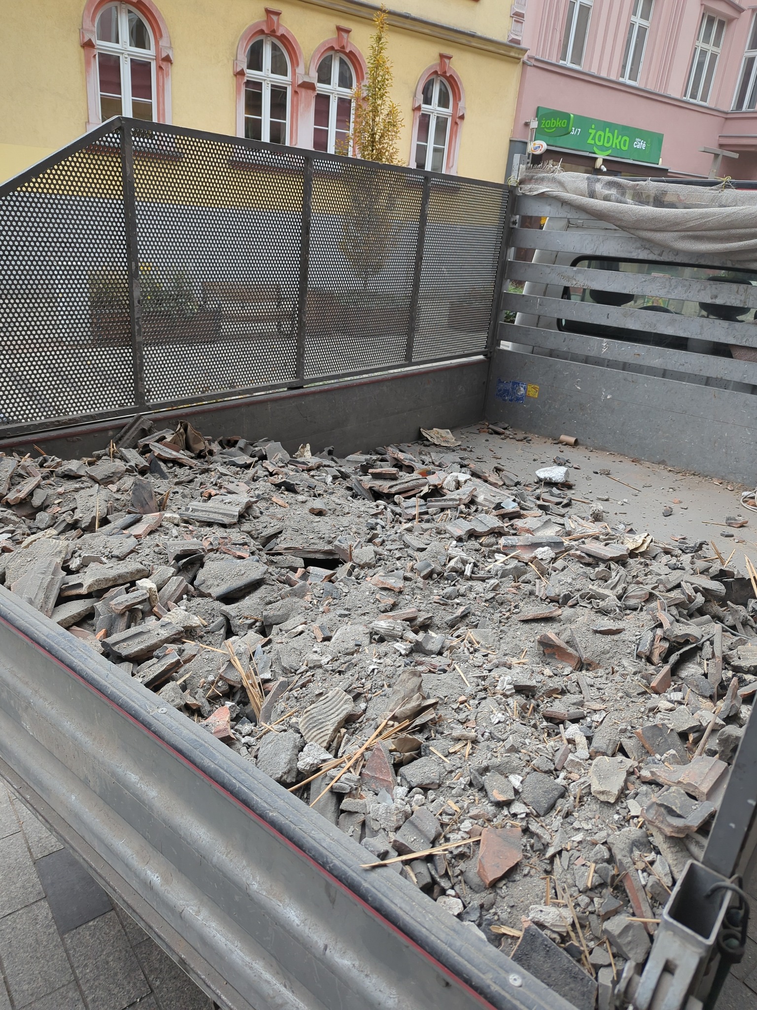 Gruz w metalowej skrzyni samochodu transportowego, widoczne fragmenty cegieł i betonu, w tle budynki miejskie z oknami łukowymi i szyld sklepu Żabka.