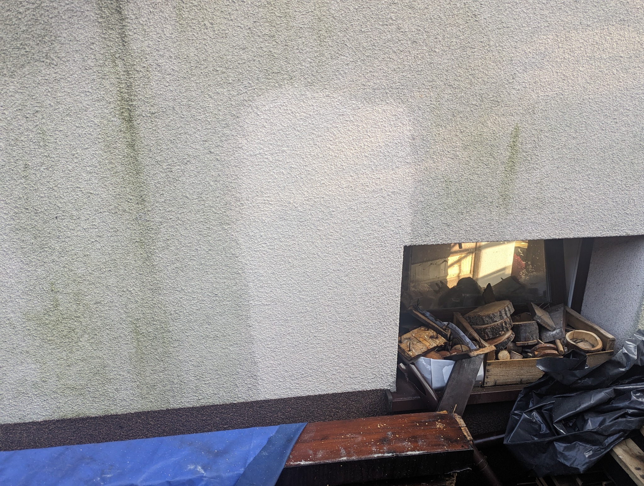 Fragment ściany budynku z widocznymi śladami zabrudzeń i wilgoci, obok otwór z drewnem opałowym i odpadami budowlanymi.