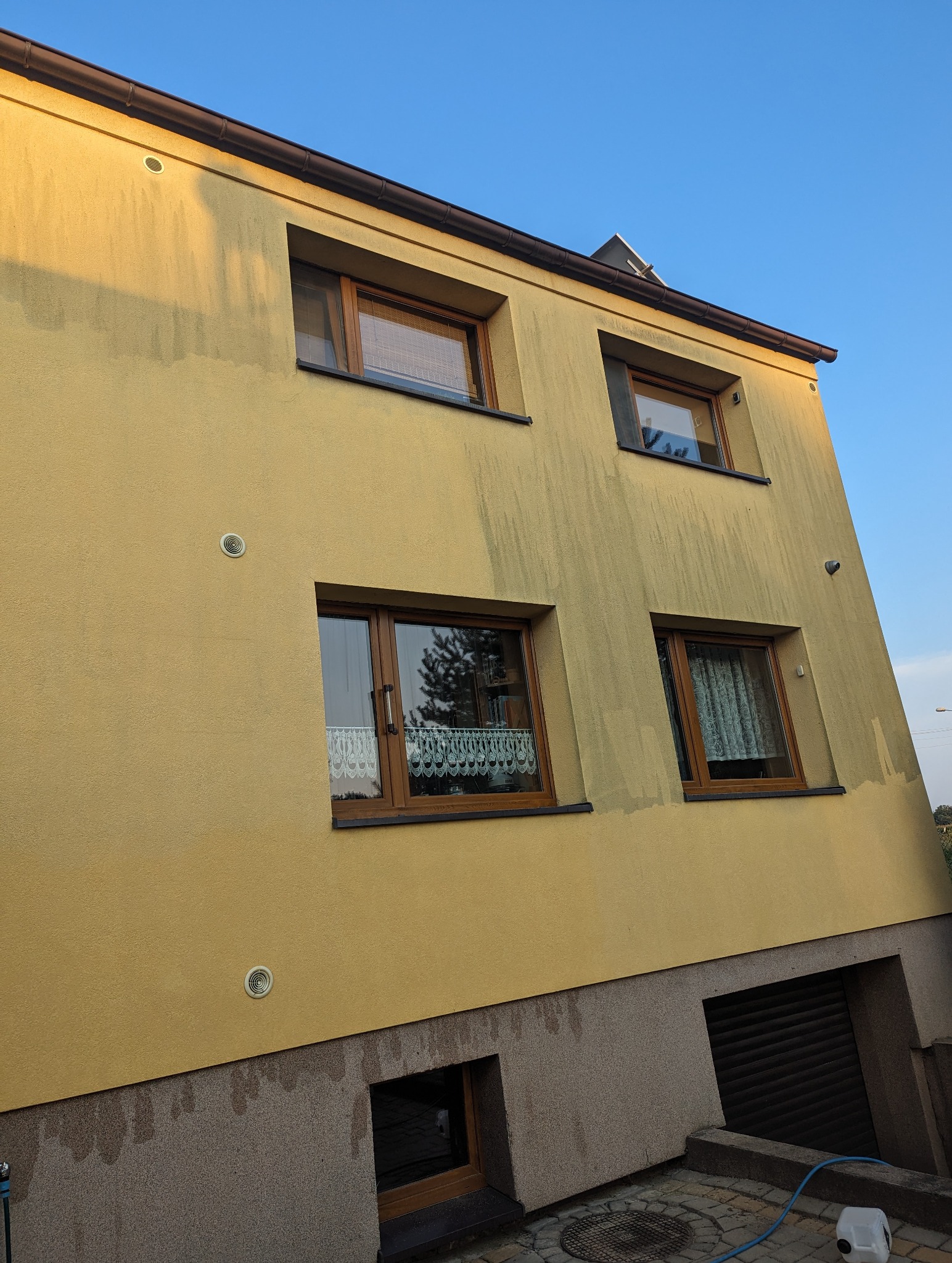 Żółta elewacja dwupiętrowego domu z brązowymi oknami, widoczne ślady po czyszczeniu, tworzące kontrast z nieoczyszczonymi fragmentami, ukazujące efekt przed i po.