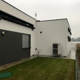 Nowoczesny dom z białą elewacją, widoczne detale architektoniczne, klimatyzator Panasonic na zewnątrz, trawnik i fragment osoby w szarej bluzie.