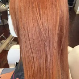 Widok z tyłu na proste włosy w odcieniu miedzianego blondu, z delikatnymi refleksami, po zabiegu koloryzacji, widoczny fragment fotela i dłoni