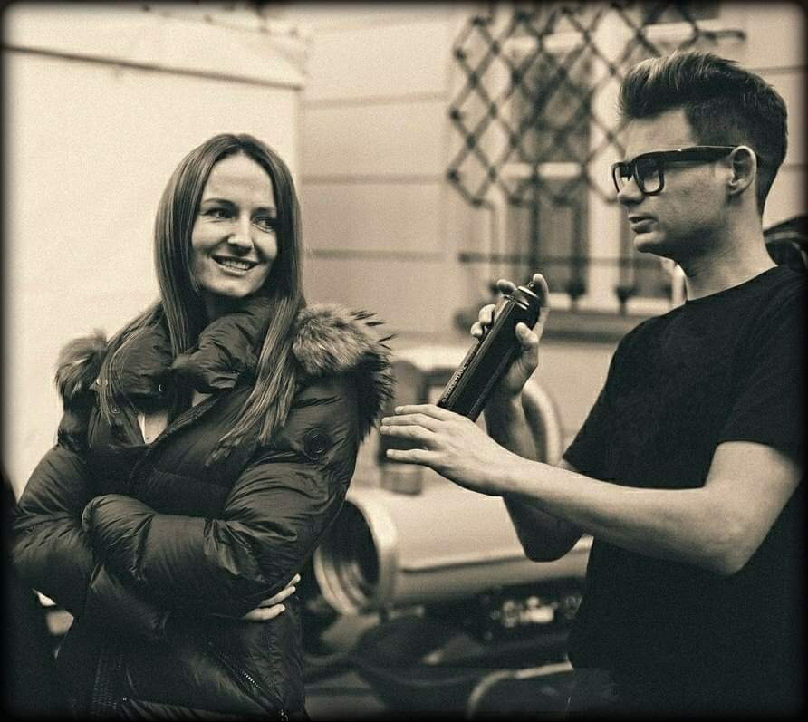 Stylista w okularach przygotowuje włosy klientki lakierem, uśmiechnięta kobieta w zimowej kurtce obserwuje proces, czarno-biała fotografia.